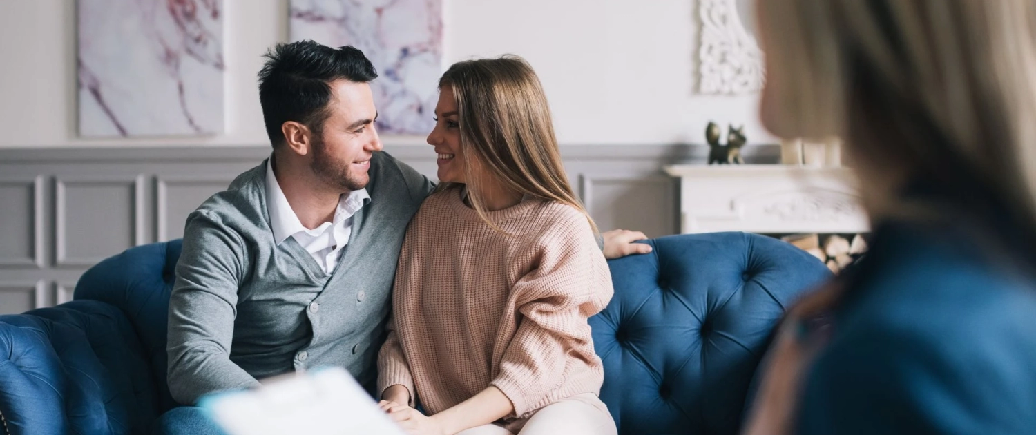 MCC-Hero-Img A couple sits closely together on a blue sofa, smiling and looking into each other's eyes. A third person, slightly out of focus and holding a clipboard, is seated in the foreground, possibly suggesting a therapy or counseling session. The room has elegant decor.