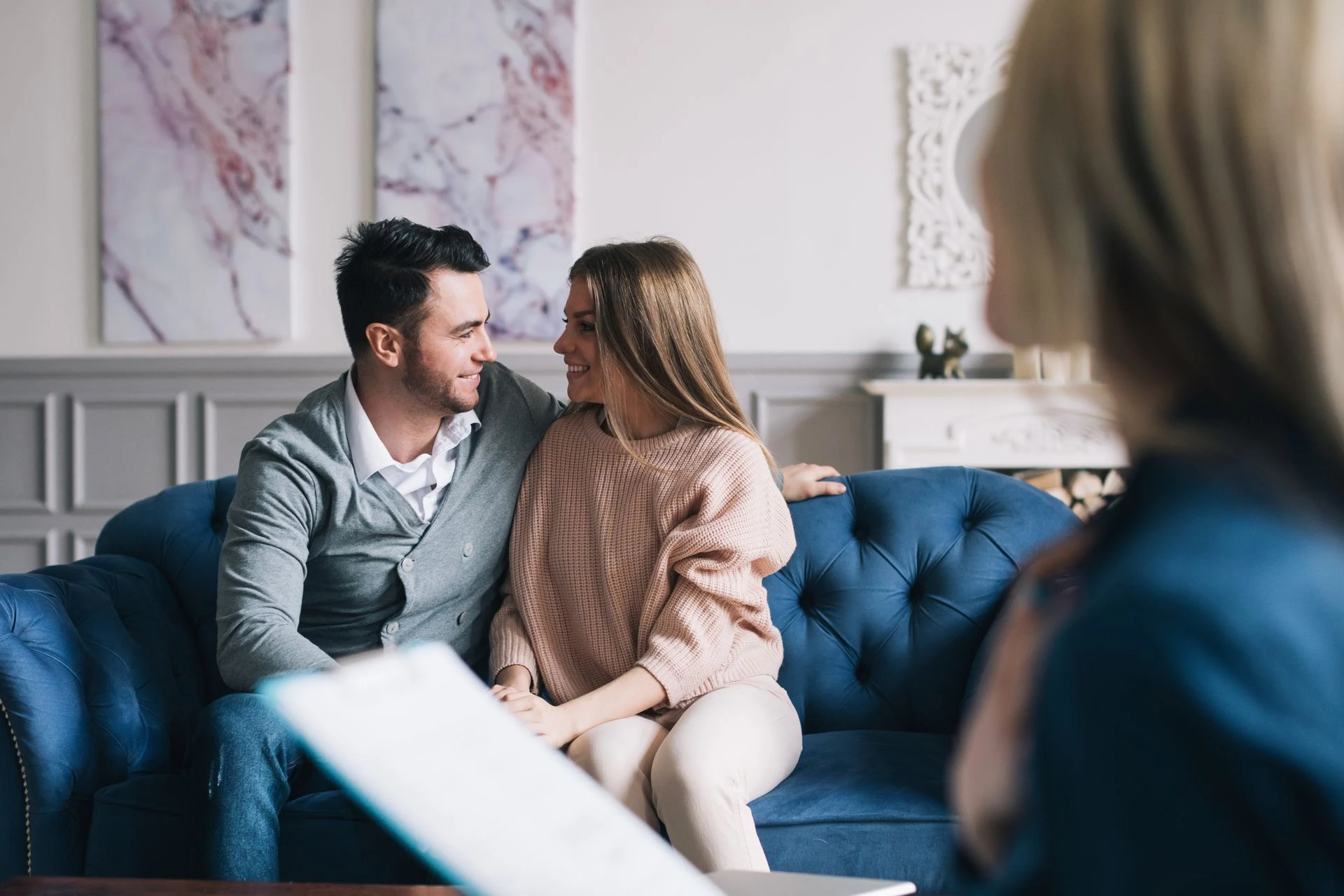 MCC-Hero-Img A couple sits closely together on a blue sofa, smiling and looking into each other's eyes. A third person, slightly out of focus and holding a clipboard, is seated in the foreground, possibly suggesting a therapy or counseling session. The room has elegant decor.